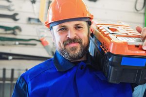 A worker in a helmet with a box of tools on his shoulder looks directly into the camera and smiles.